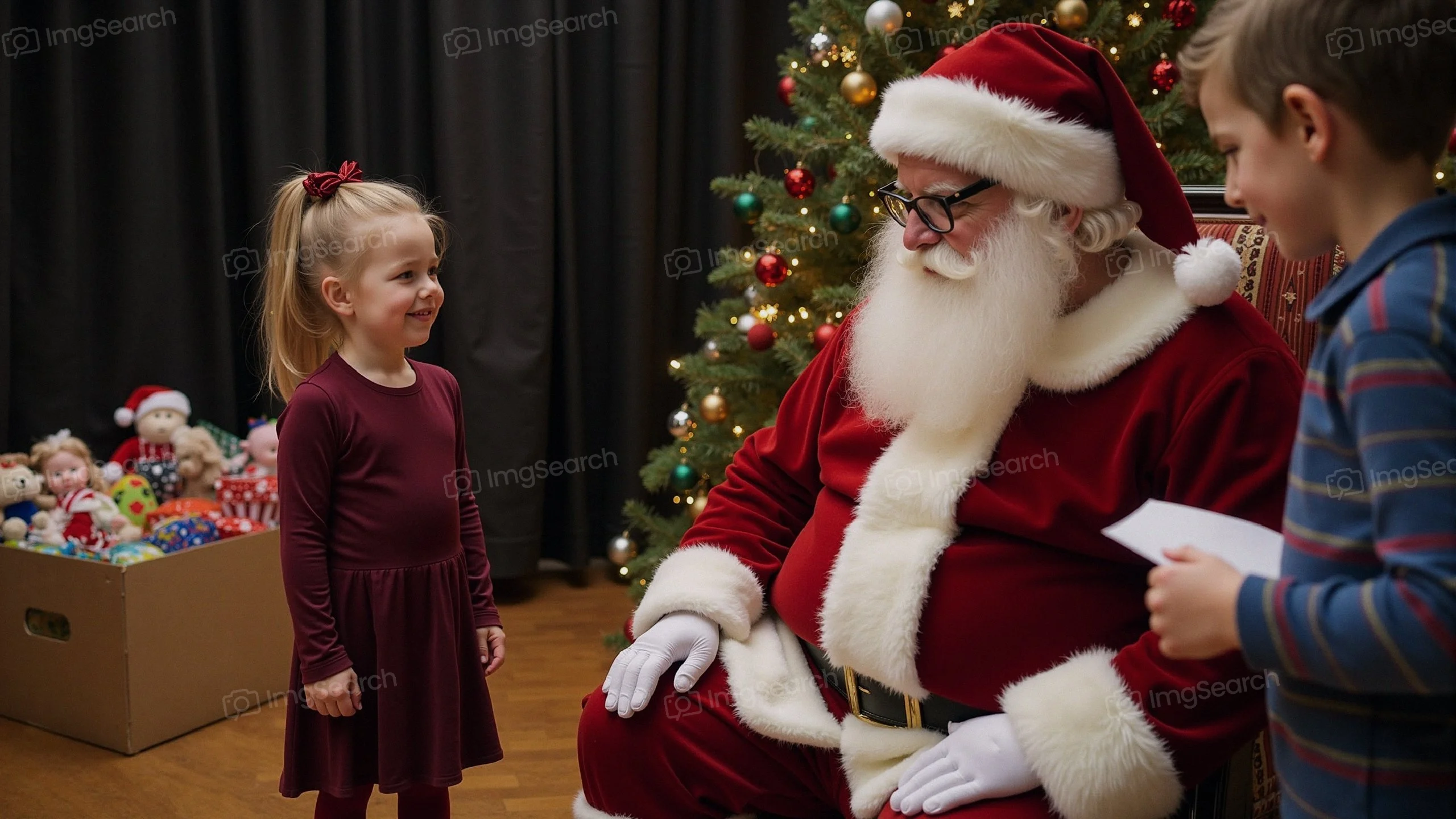 A young girl in a burgundy dress talking to Santa Claus in a festive room with a Christmas tree.
