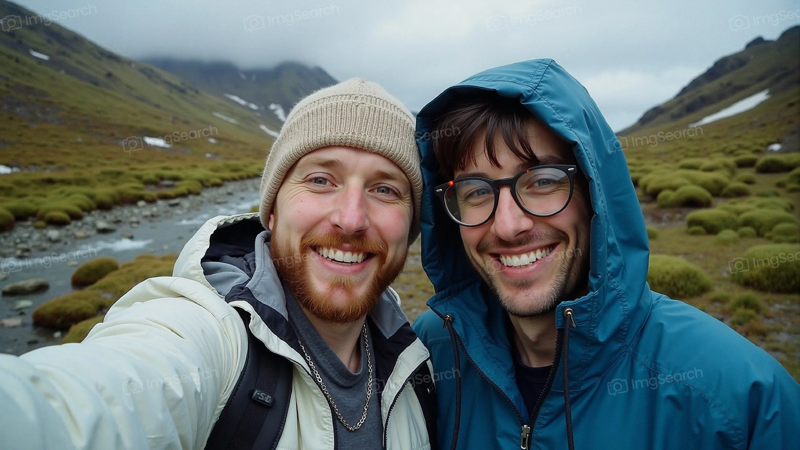 Two friends smiling for a selfie in a green mountainous landscape under a cloudy sky.