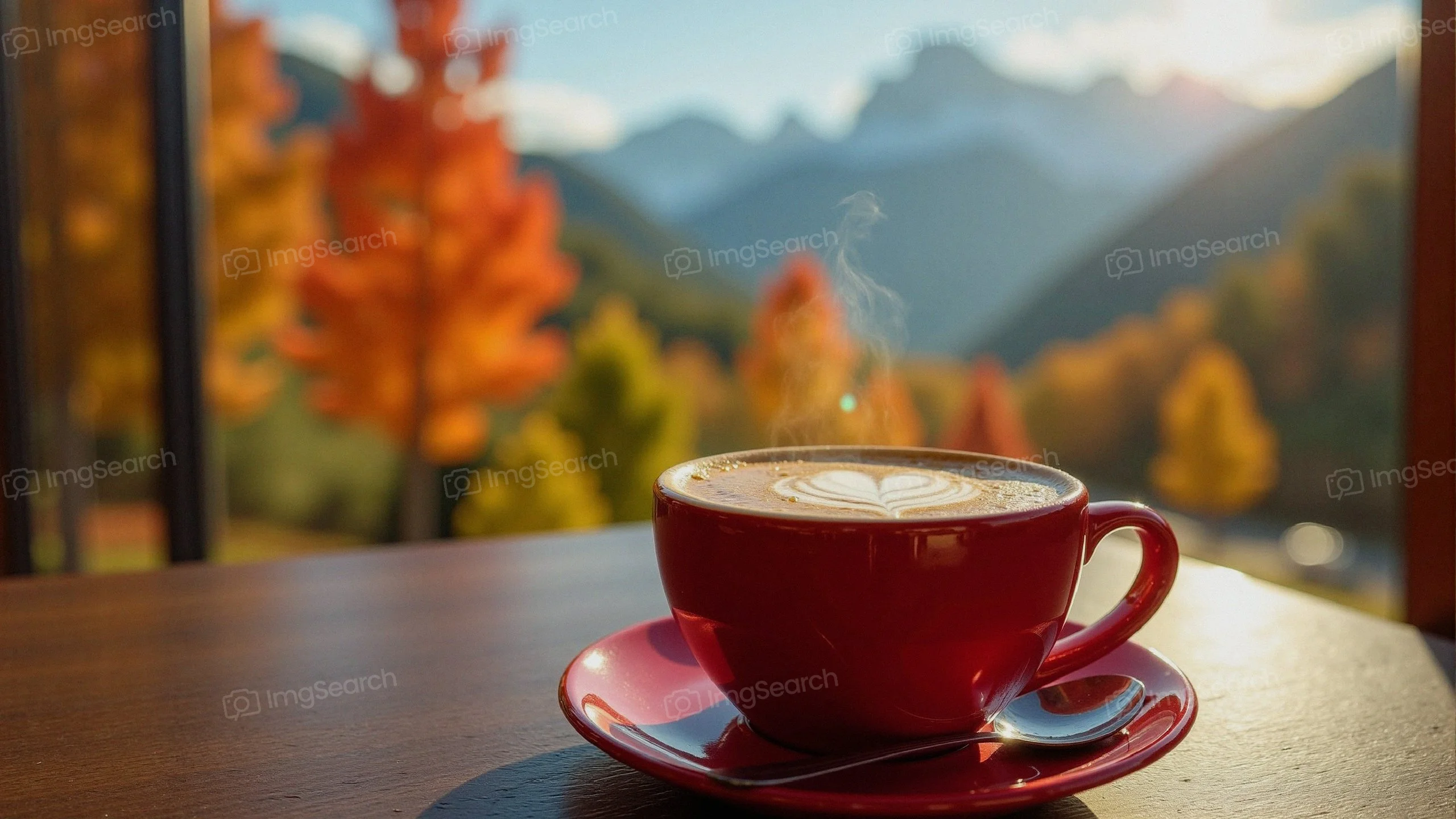 A red cup of coffee with latte art on a saucer in front of a mountain landscape with autumn trees.