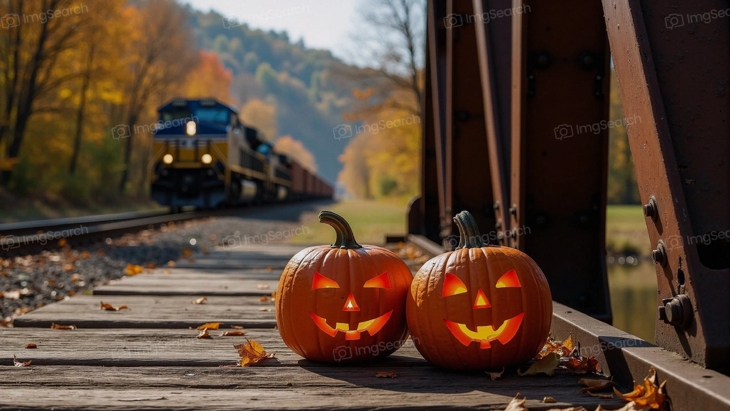 Two glow-in-the-dark jack-o'-lanterns on a wooden train track with autumn trees a train in the background.