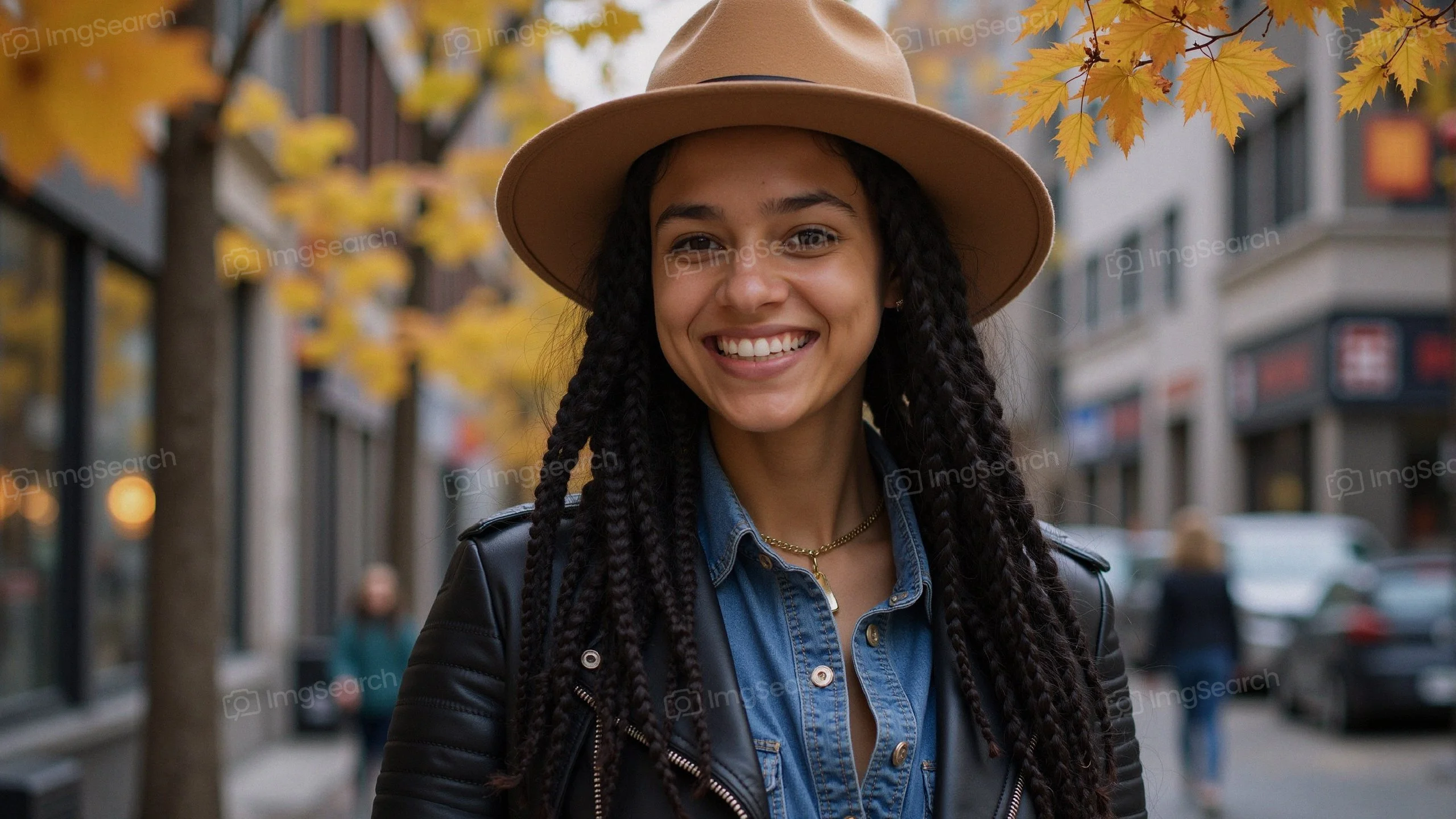 A young woman with braids a brown hat smiling in an autumn setting with golden leaves.