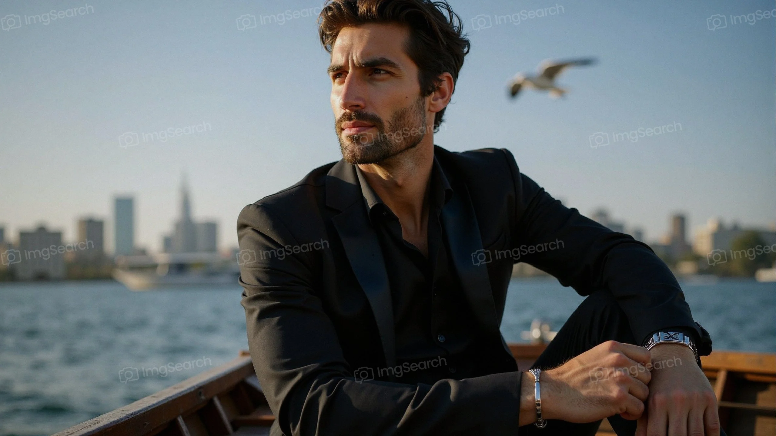 A man in a black suit sitting on a boat with a city skyline in the background a seagull flying overhead.