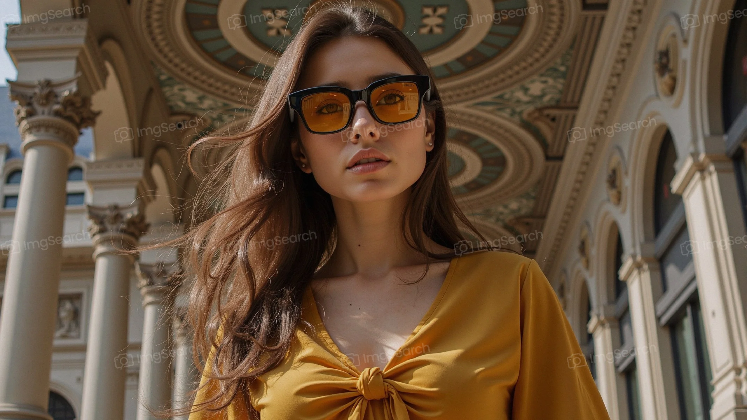 A young woman in a yellow blouse sunglasses stands under an ornate archway, with flowing hair a confident expression.