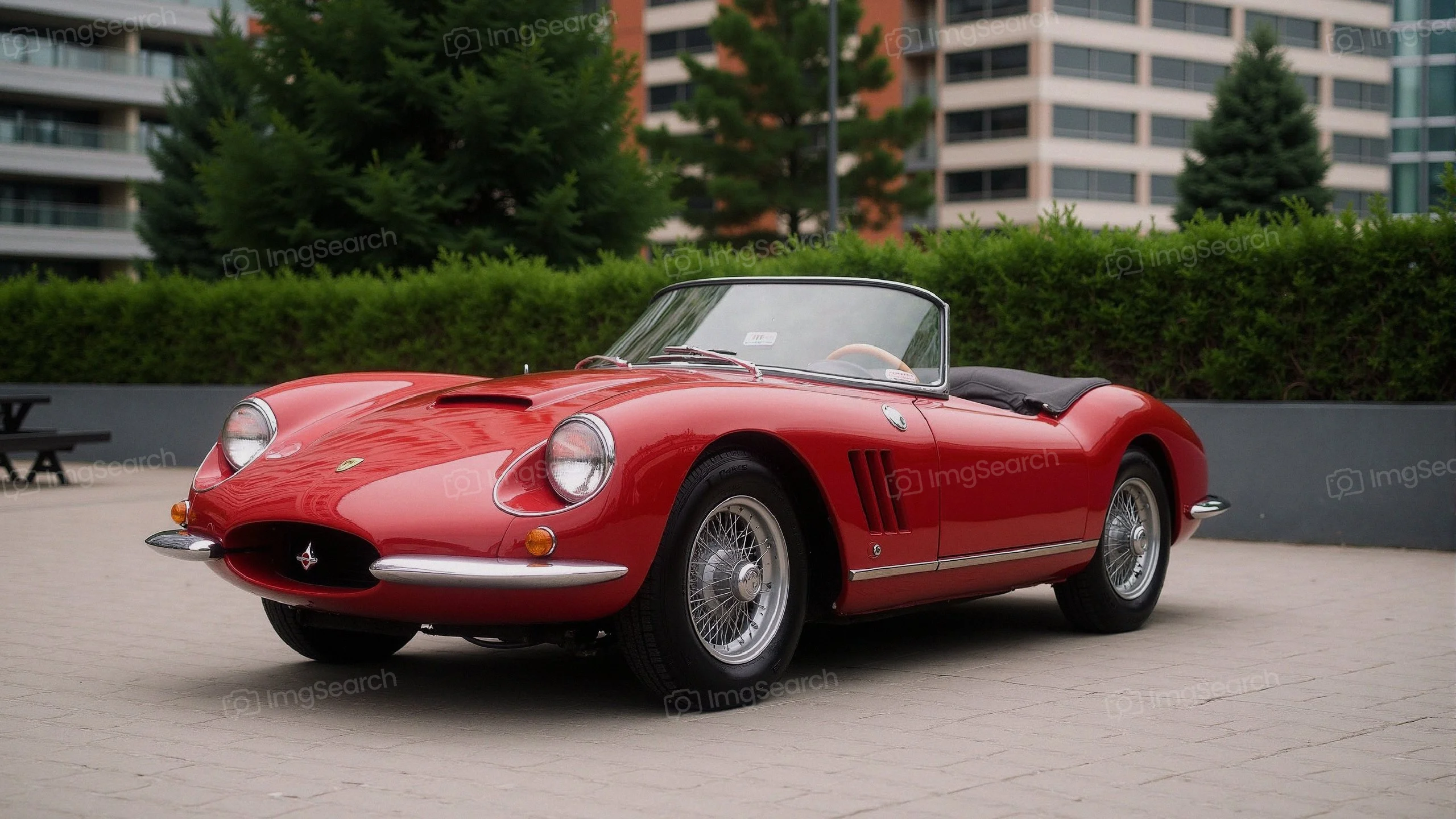 A classic red sports car parked in an urban area with greenery in the background.