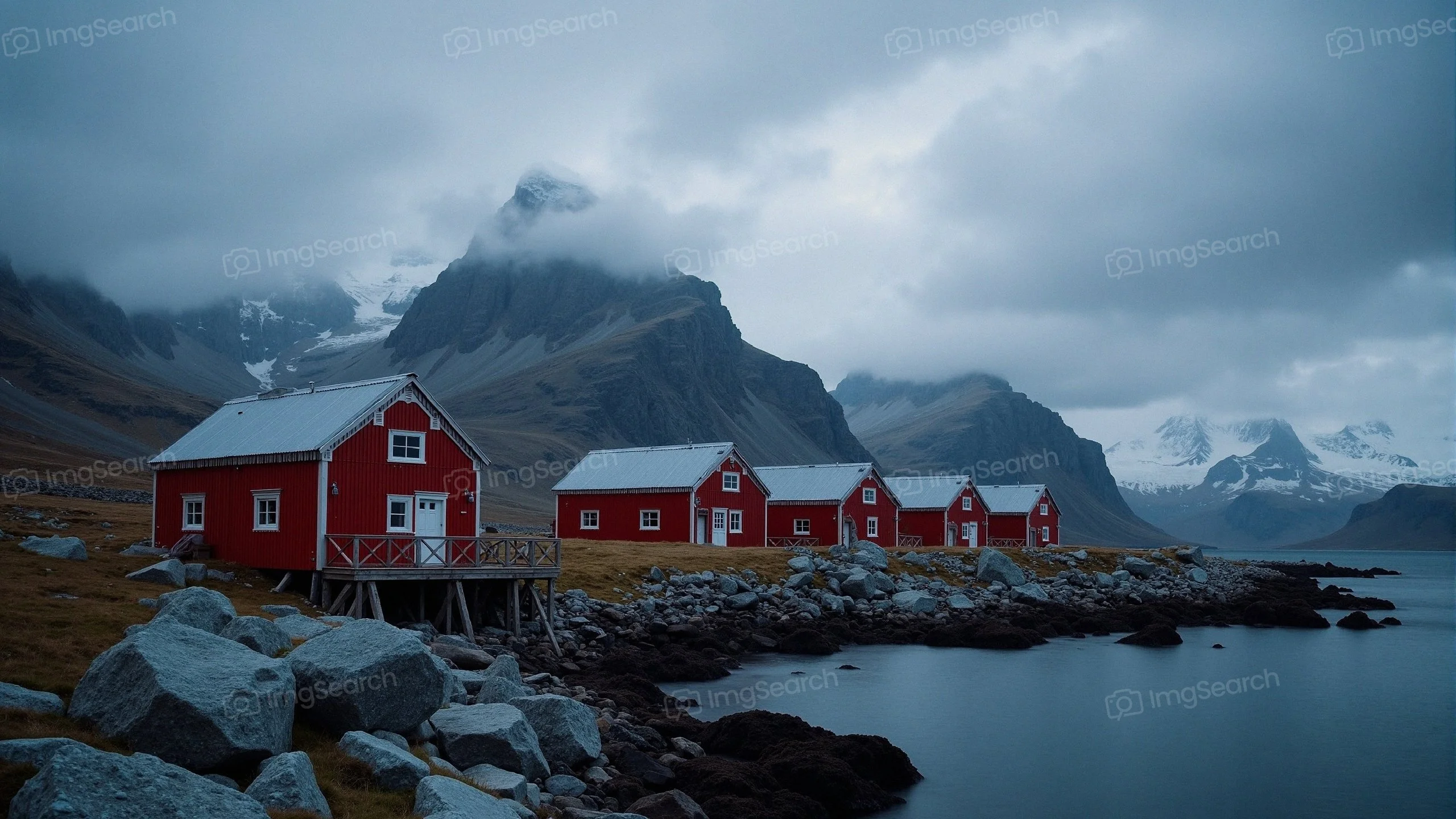 A row of red wooden houses by the calm waters with mountains in the background under a cloudy sky.