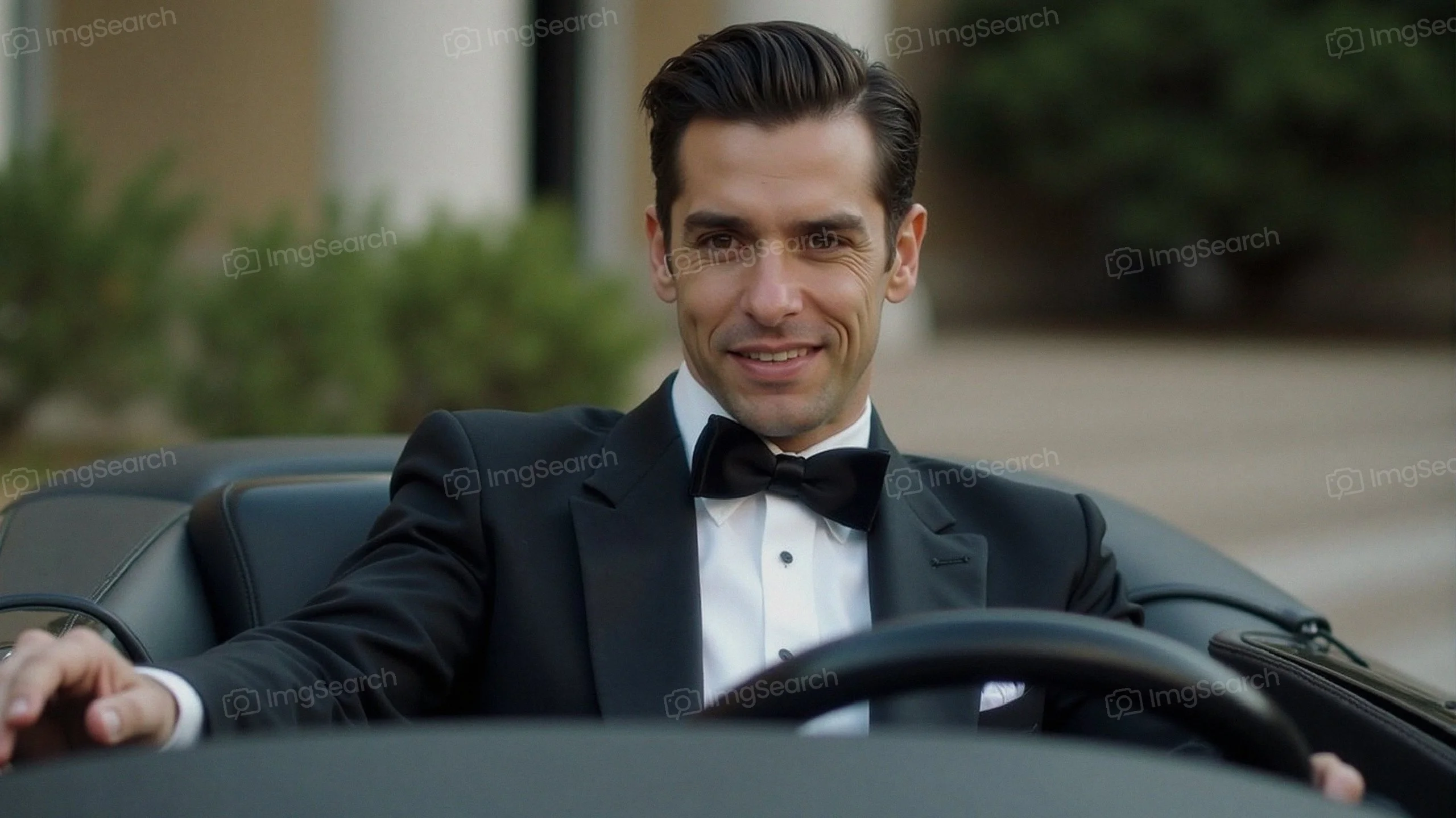 A man in a tuxedo sits confidently in the driver's seat of a classic convertible car.