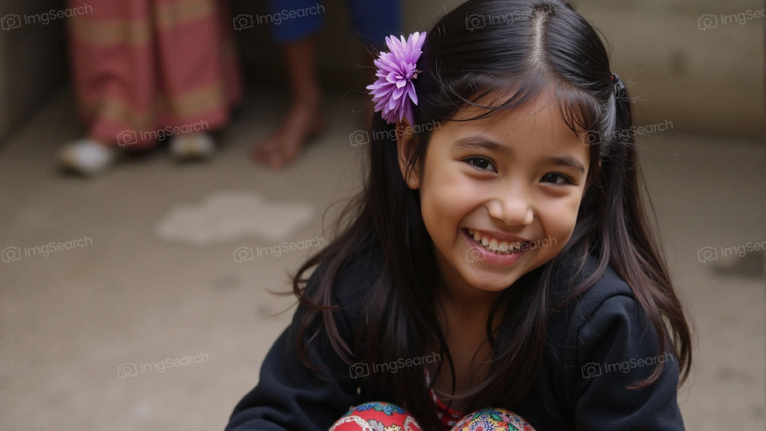 A young girl smiling brightly, wearing a purple flower in her hair.