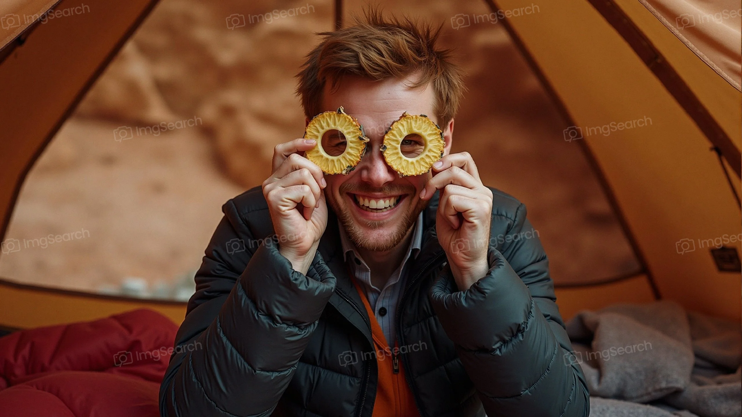 A man holding pineapple rings in front of his eyes while inside a camping tent, smiling joyfully.