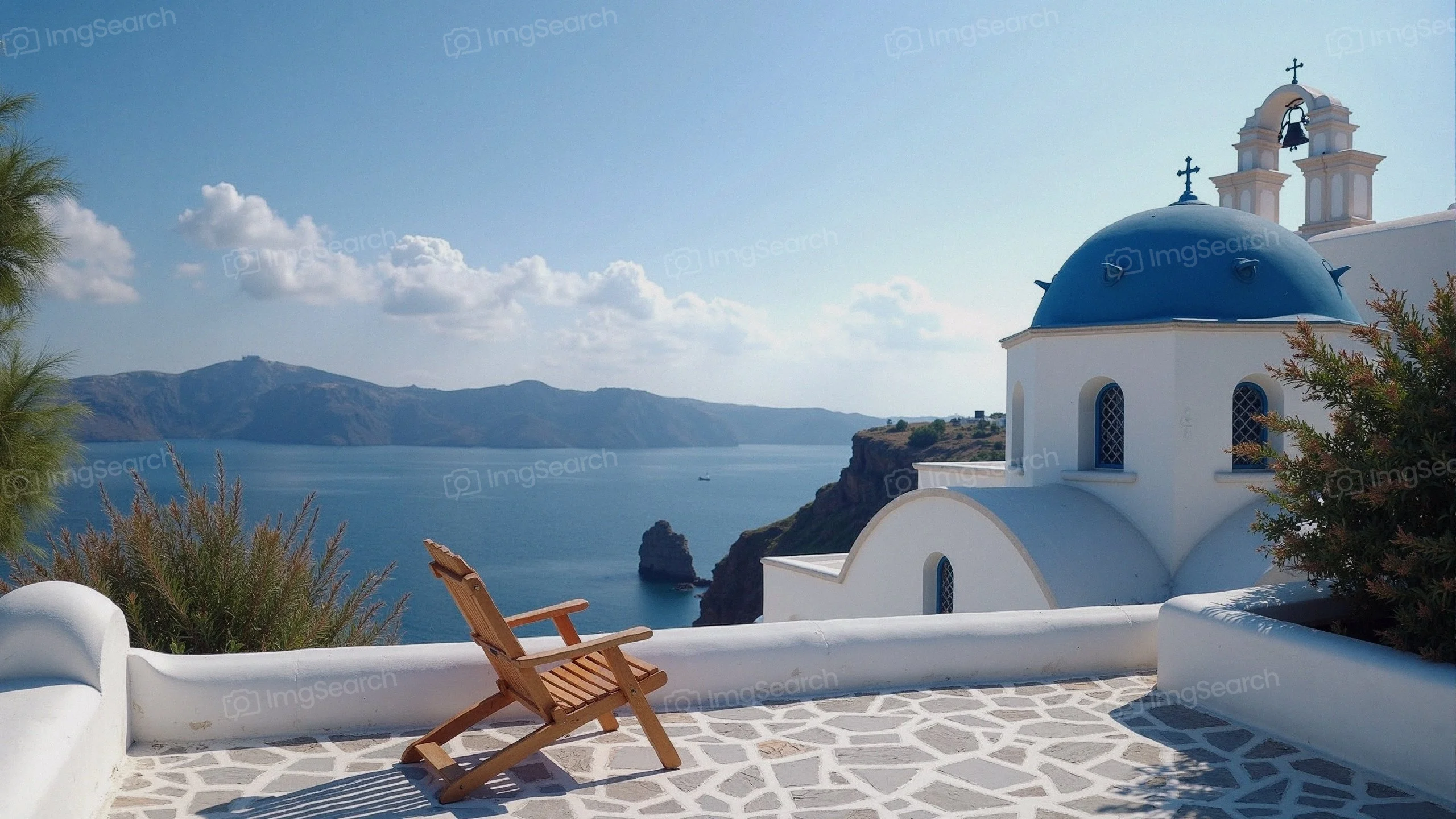 A wooden lounge chair on a stone terrace overlooking the Aegean Sea with blue-domed buildings in Santorini.