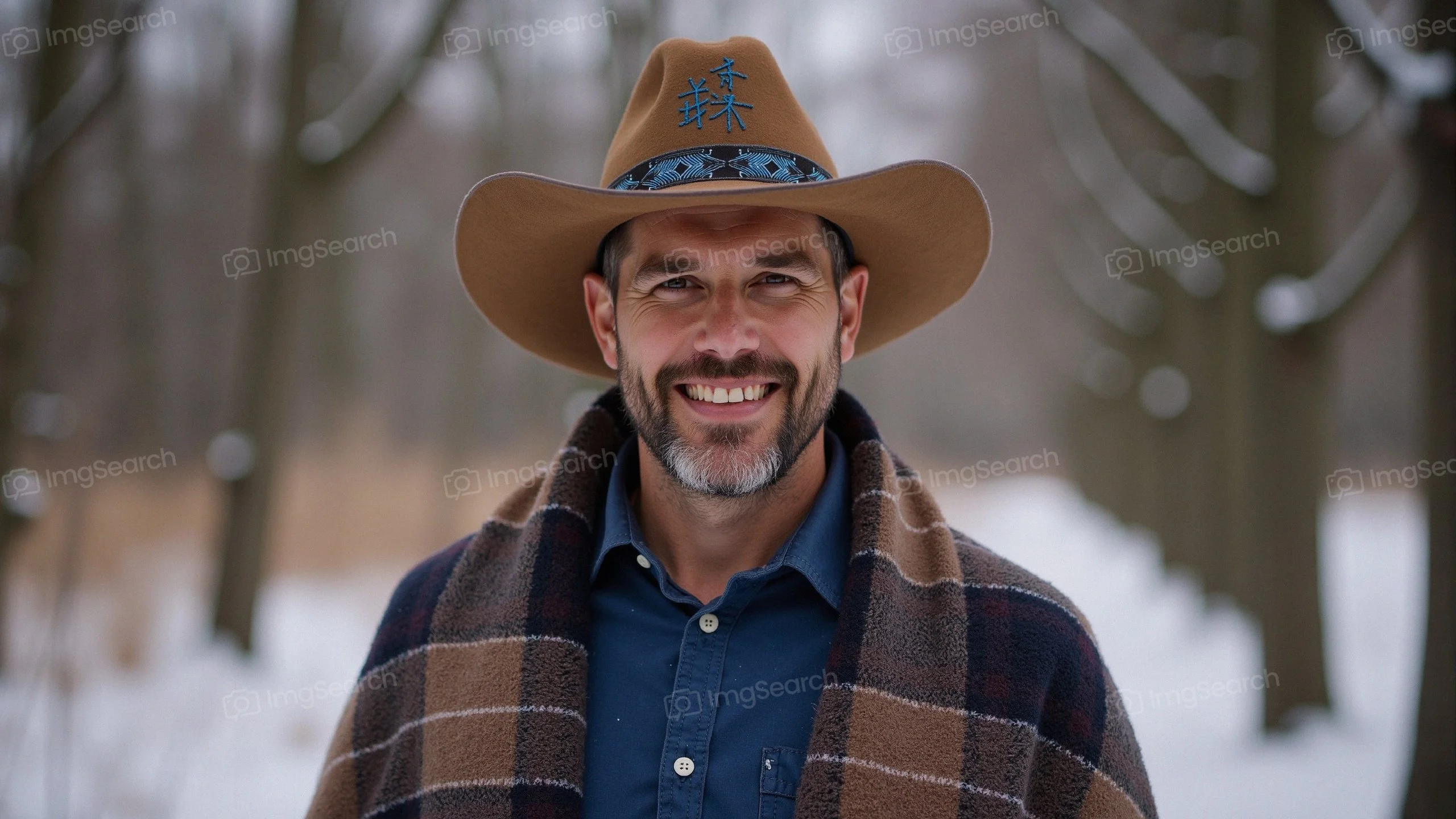 A smiling man in a cowboy hat plaid blanket standing in a snowy landscape