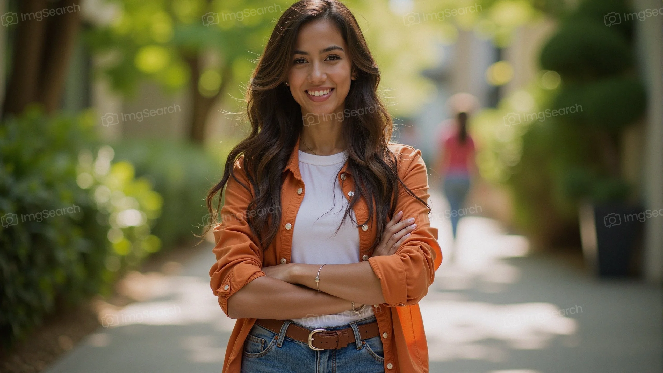 A smiling young woman with long hair wearing an orange shirt jeans stands in a green outdoor pathway.
