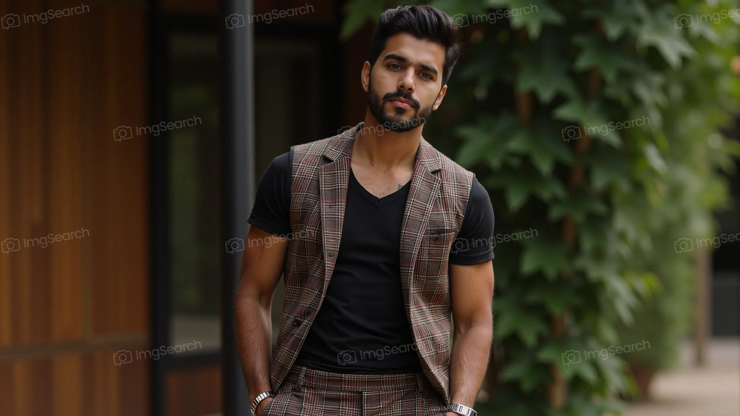 A stylish young man in a plaid blazer black t-shirt stands in front of greenery.