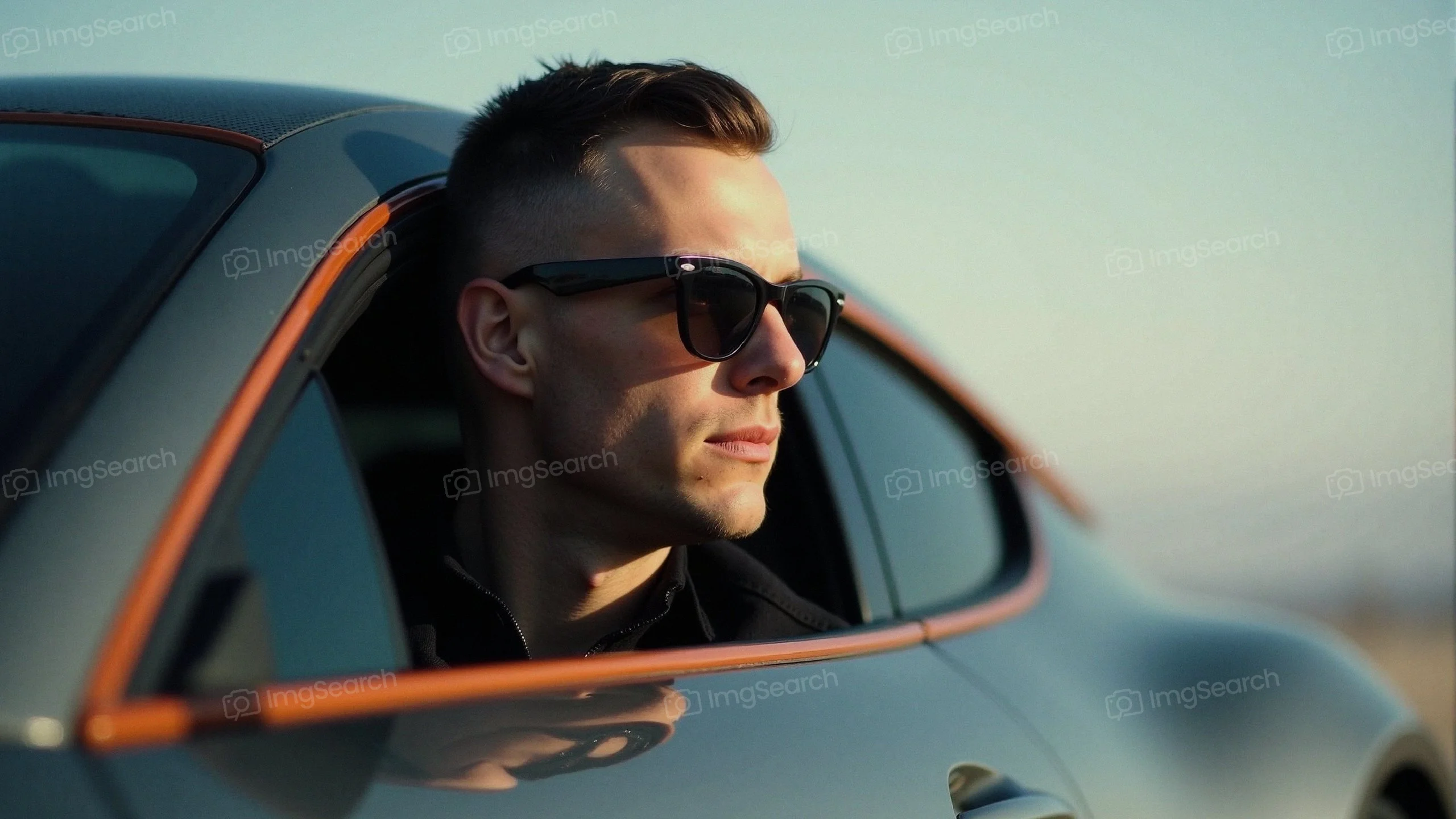 A young man wearing sunglasses leans out of a sleek sports car.