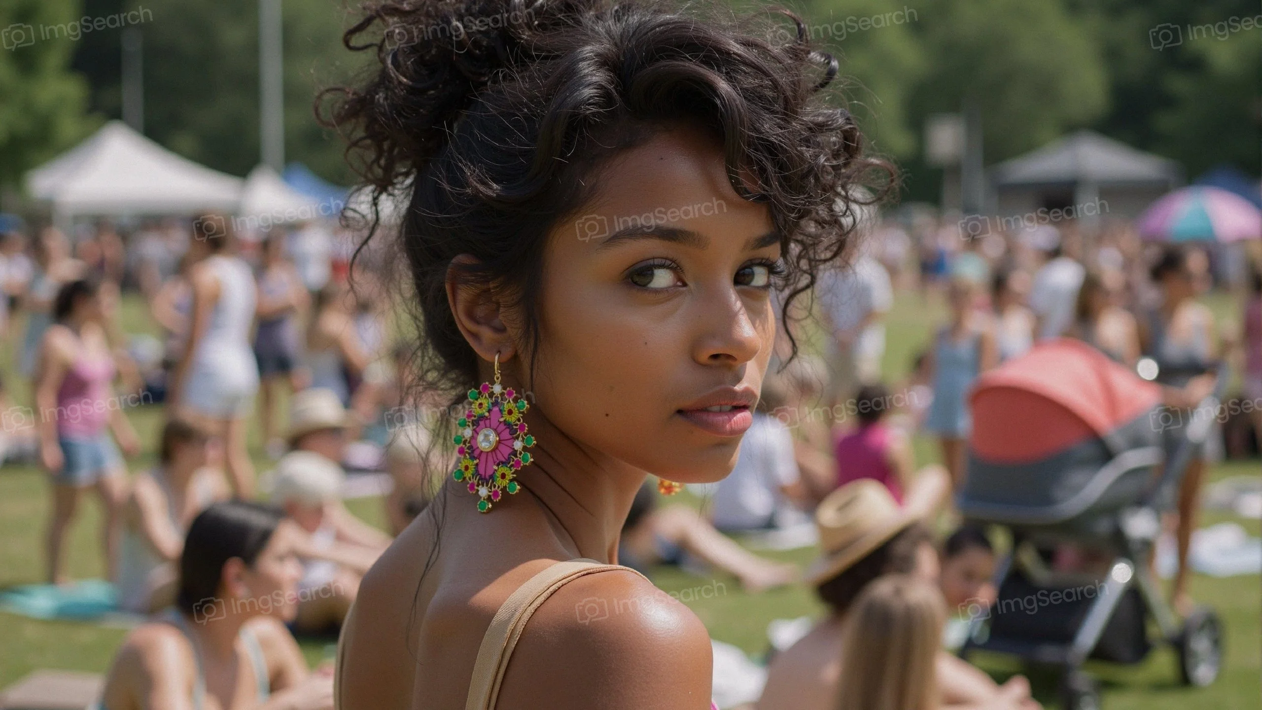 A young woman with colorful earrings looking back at the camera in a bustling outdoor festival scene.