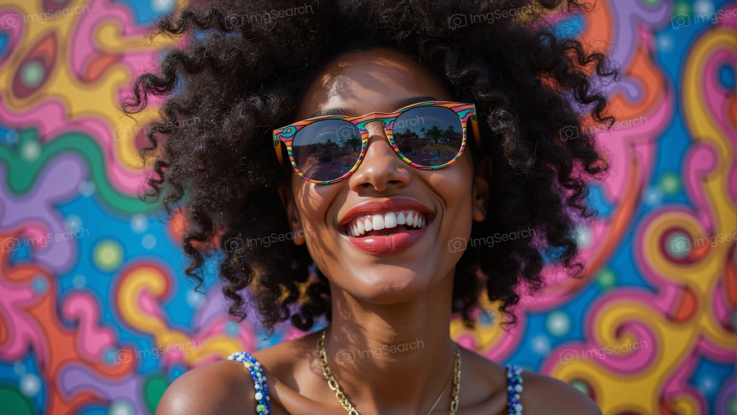 A young woman with curly hair smiling, wearing sunglasses in front of a colorful mural.