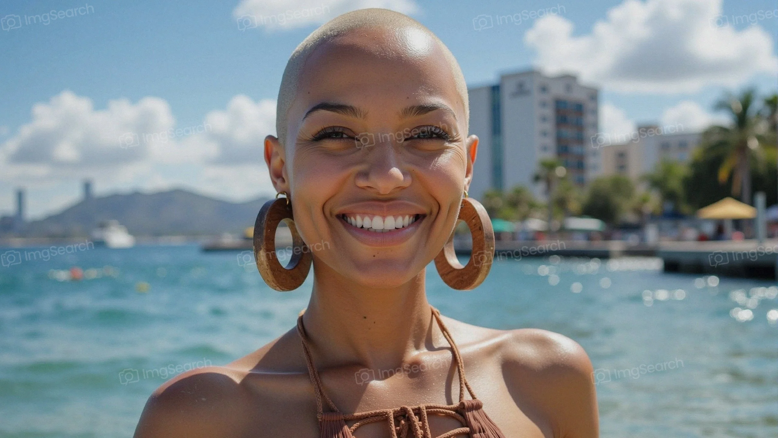 A smiling woman with a shaved head wooden hoop earrings standing by the beach on a sunny day.