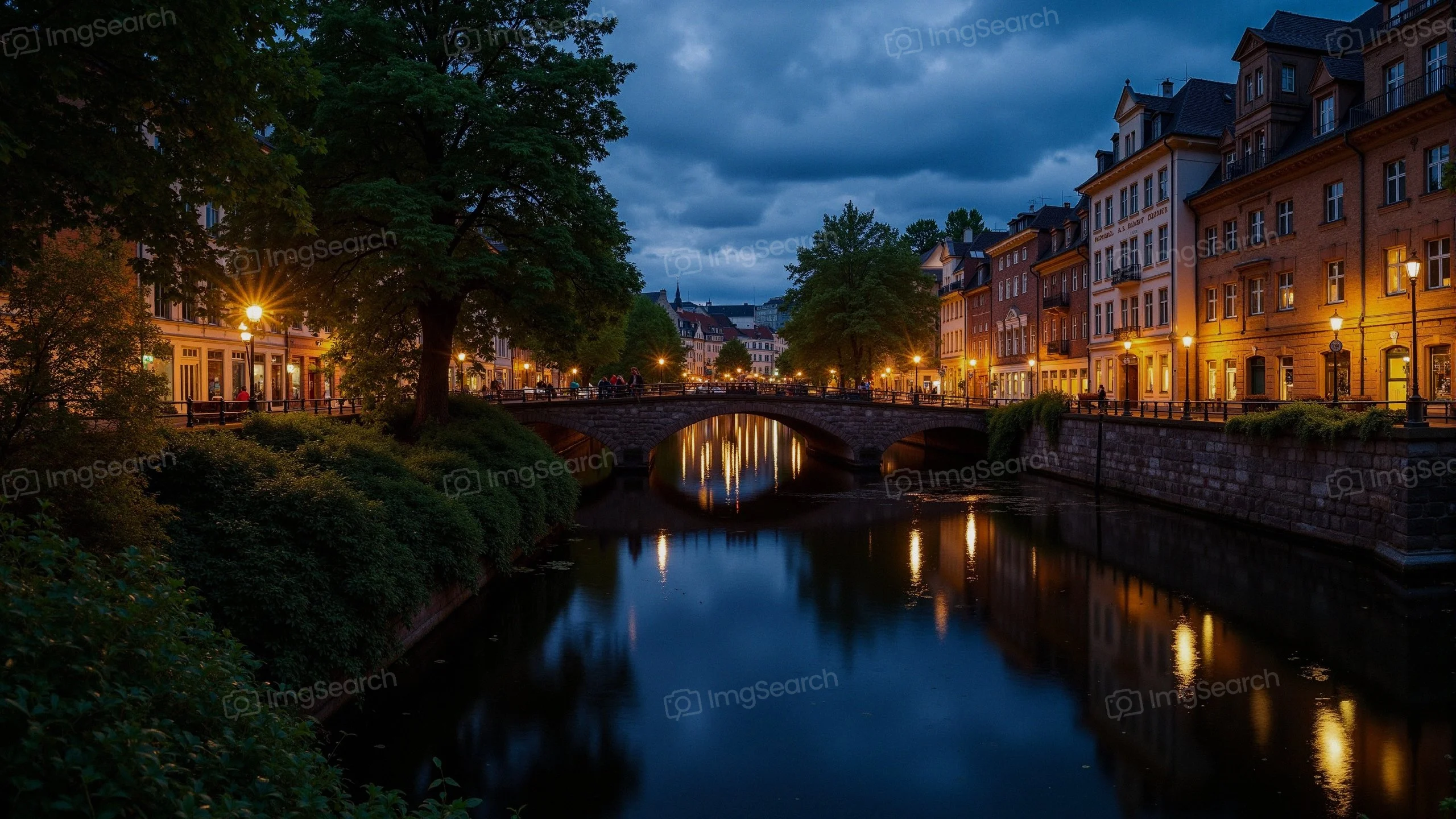 A picturesque view of a stone bridge over a calm river, illuminated by soft golden lights from surrounding buildings trees at dusk.