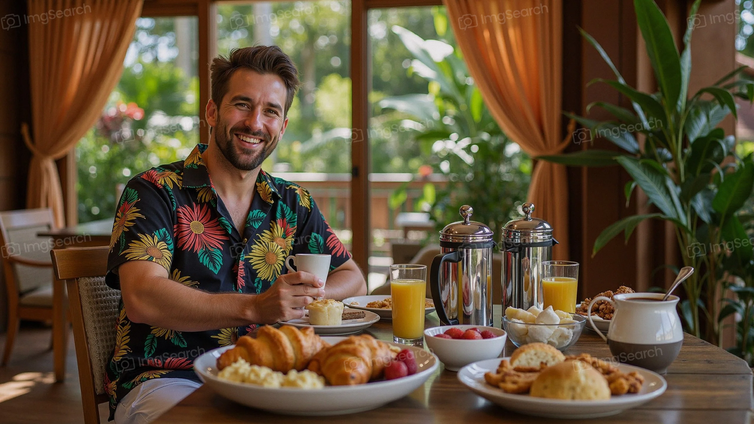 A man in a floral shirt enjoying a tropical breakfast with croissants, fruits, drinks in a sunny room.
