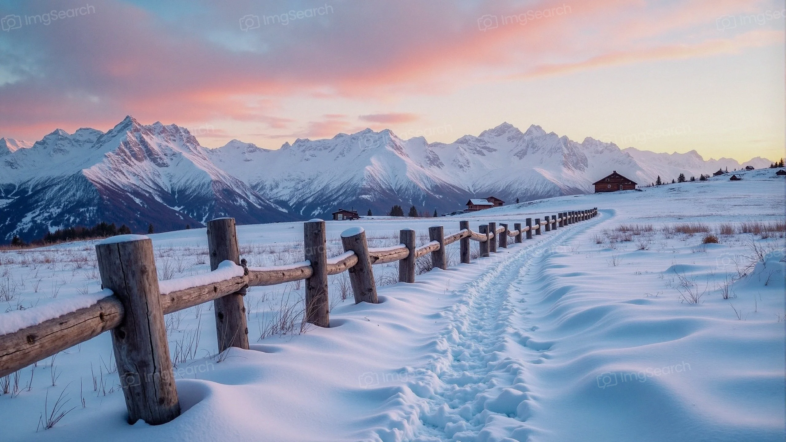 A snowy landscape at dusk with mountains in the background a wooden fence along a path.