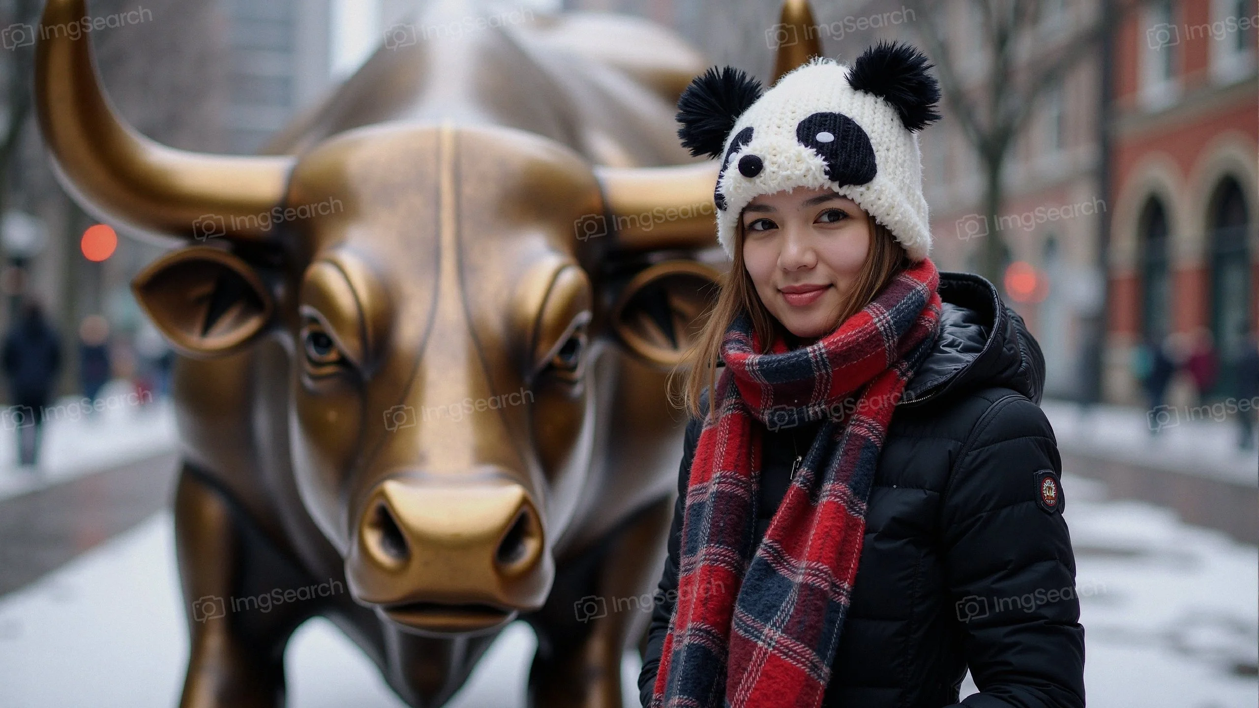 A woman in a panda hat plaid scarf stands smiling in front of a golden bull statue in a snowy scene.