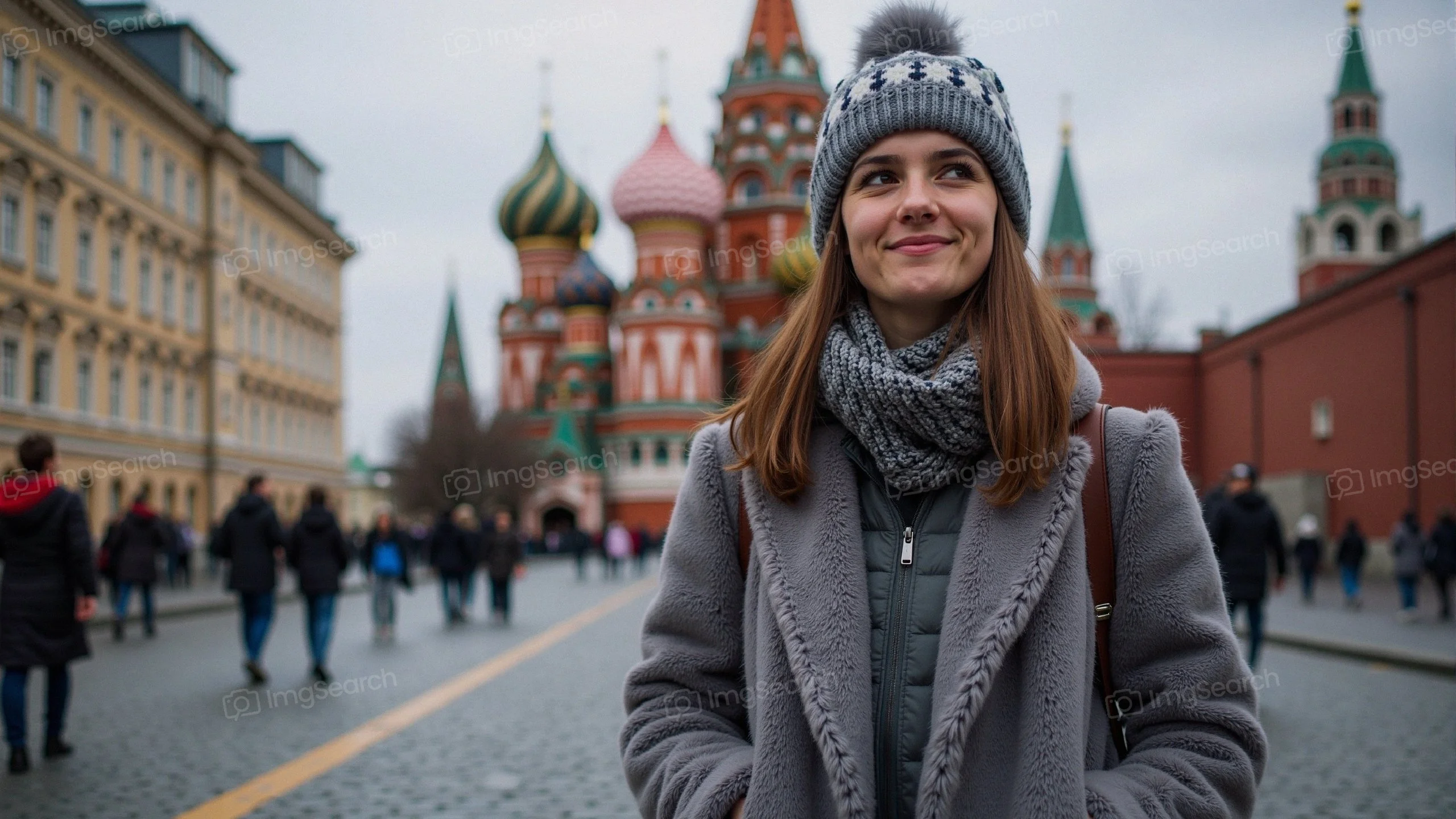 A young woman wearing a gray coat beanie stands in front of St. Basil's Cathedral in Moscow, with people walking in the background.